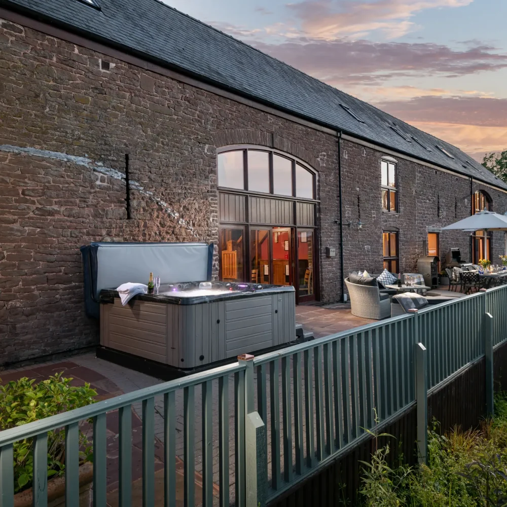Raised patio at dusk with outdoor shower, hot tub, comfortable day bed and outdoor dining, Penblaith Barn
